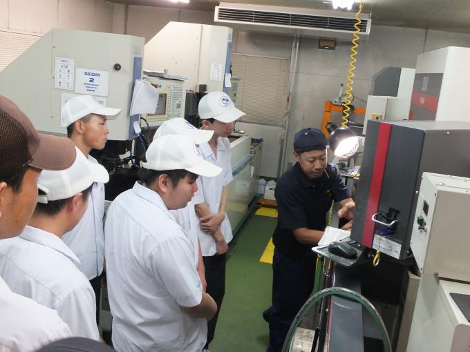 工場で機械を動かしながら説明する作業員と、話を聞く生徒達の写真