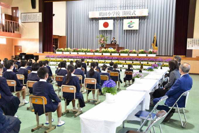 新鶴田小学校の開校式・入学式にて、壇上の男性の話を座って聞く小学生と来賓者の写真
