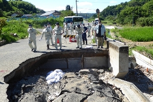 作業着を着用したさつま町議員達が豪雨にて陥没した道路の災害調査を行っている写真