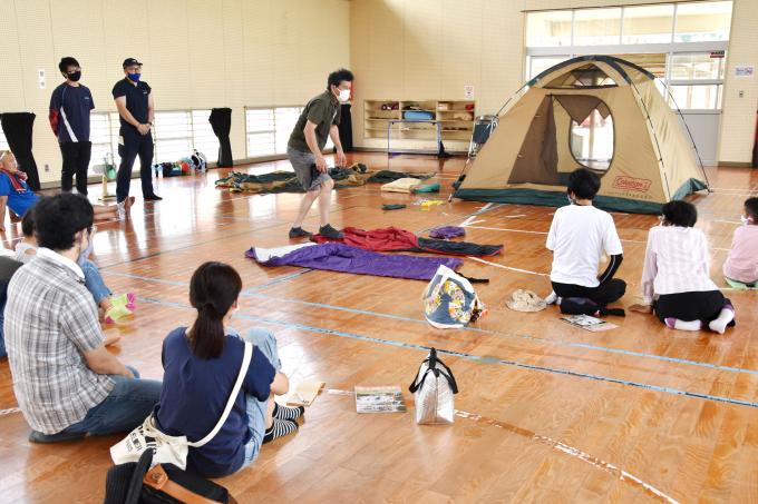 体育館でキャンプの張り方を説明する男性と、話を聞く参加者の写真