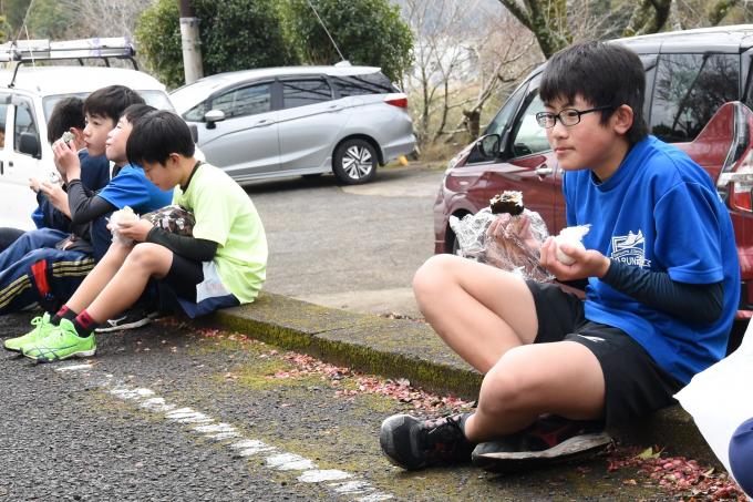 沿道に座っておにぎりを食べる小学生の写真