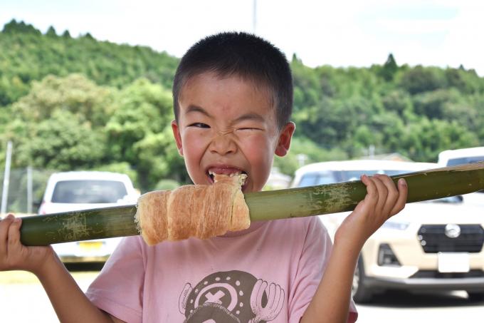 竹に巻き付いた焼きたてのパンをかじる男の子の写真