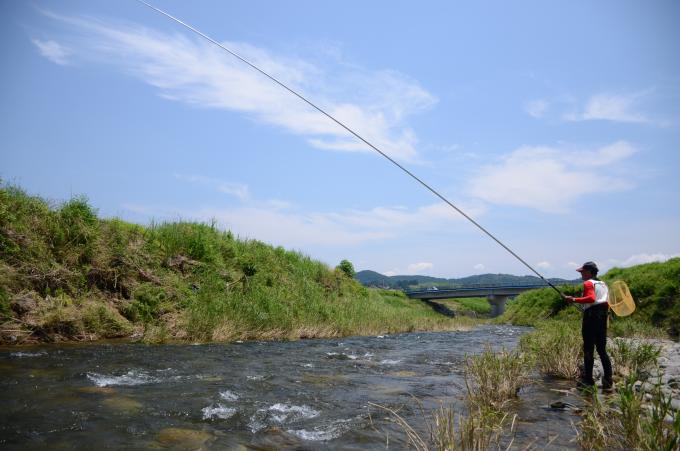 カゴを背負って河原から川に釣竿を垂らす人の写真