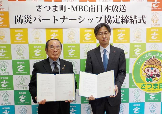 さつま町・MBC南日本放送防災パートナーシップ協定締結式と書かれた垂れ幕の前で締結書を持つ、さつま町長とMBC南日本放送取締役の写真
